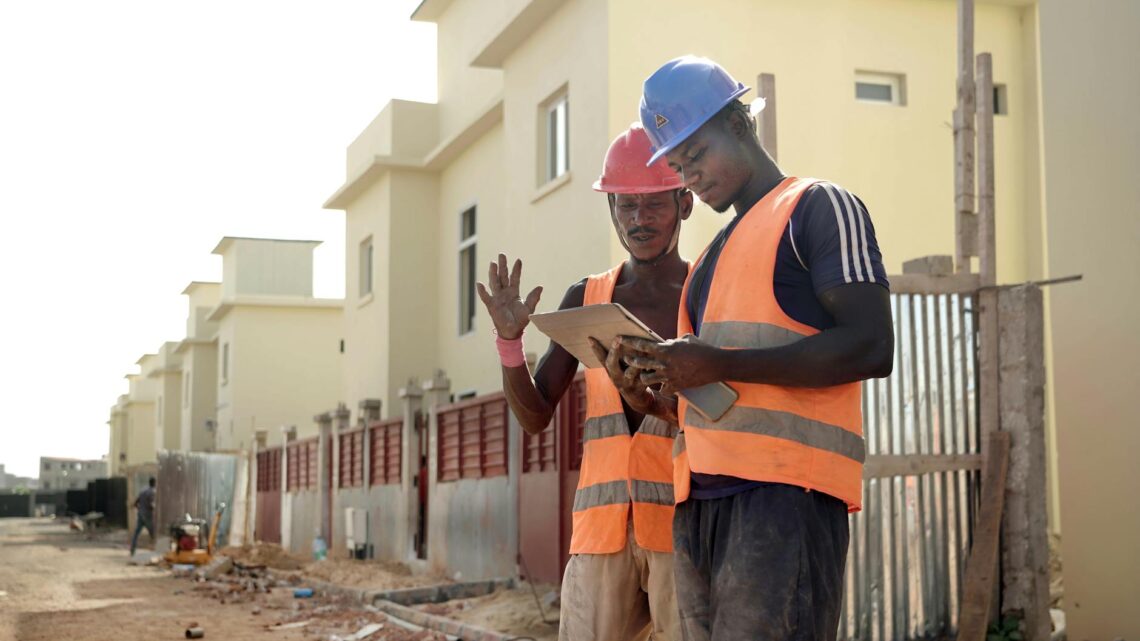 Two construction workers in helmets and vests review blueprints on a site.