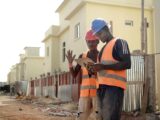 Two construction workers in helmets and vests review blueprints on a site.