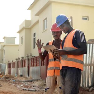 Two construction workers in helmets and vests review blueprints on a site.