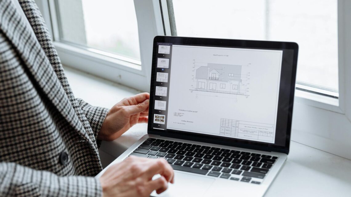 Person in plaid blazer reviewing architectural blueprints on laptop by a window.