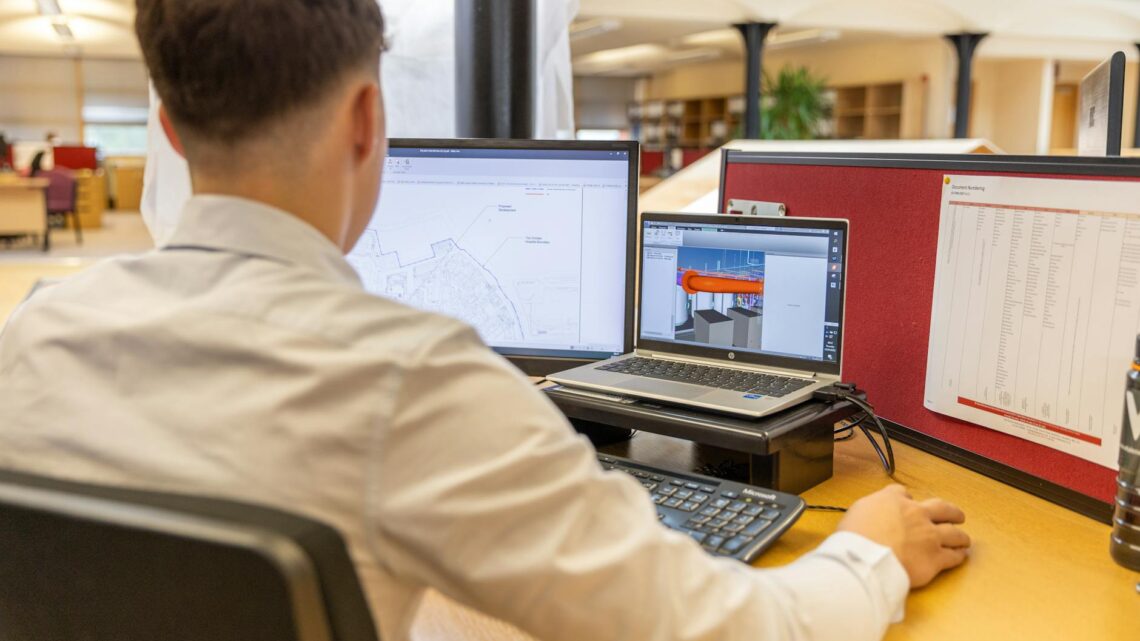Back view of engineer focused on CAD design at dual monitors in bright office setting.