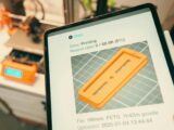 Close-up of a digital tablet displaying a 3D printing process in a workshop setting.