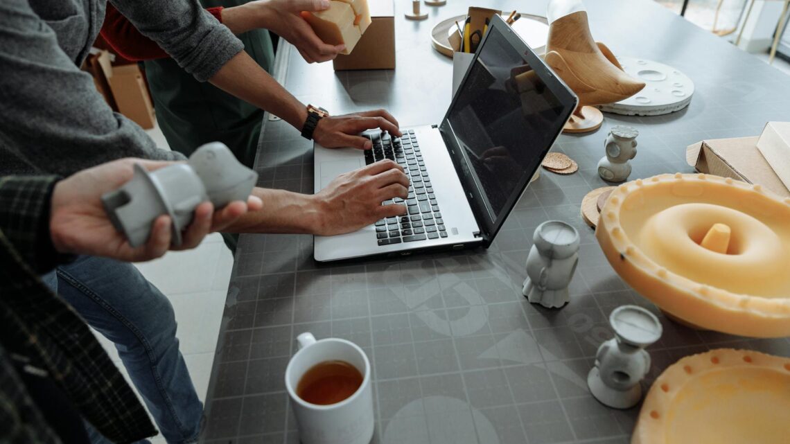 Hands on laptop in a creative workspace with 3D models and prototypes.