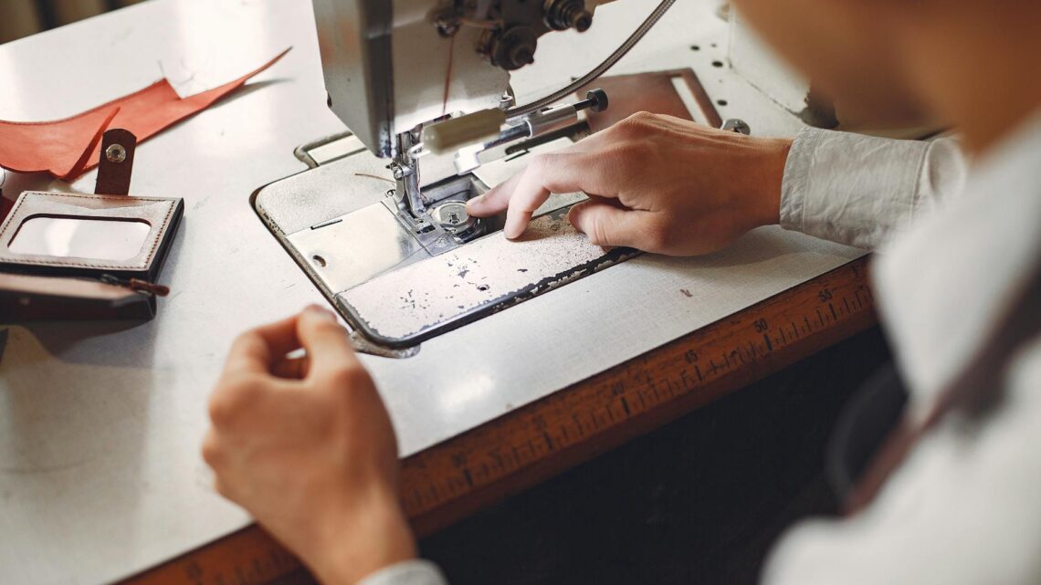 From behind crop unrecognizable artisan in white shirt sewing handmade leather details while sitting at table with sewing machine and leather wallet in daytime
