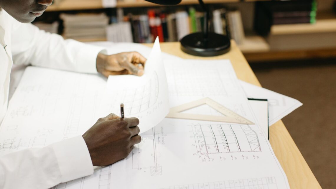 Black male architect working on blueprints at a desk in a well-lit office.