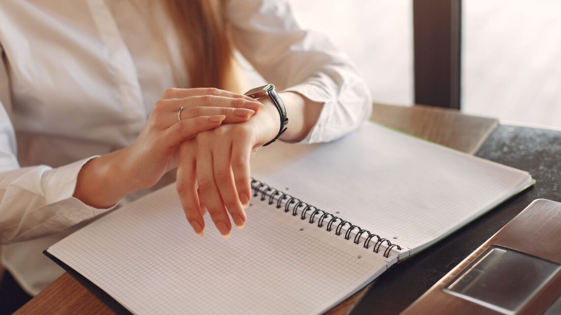 Unrecognizable crop female entrepreneur in formal clothes sitting at wooden table near laptop with copybook and checking time on wristwatch