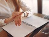 Unrecognizable crop female entrepreneur in formal clothes sitting at wooden table near laptop with copybook and checking time on wristwatch