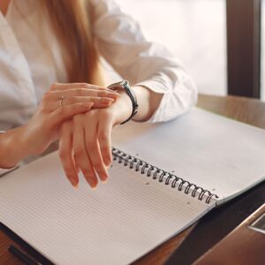 Unrecognizable crop female entrepreneur in formal clothes sitting at wooden table near laptop with copybook and checking time on wristwatch