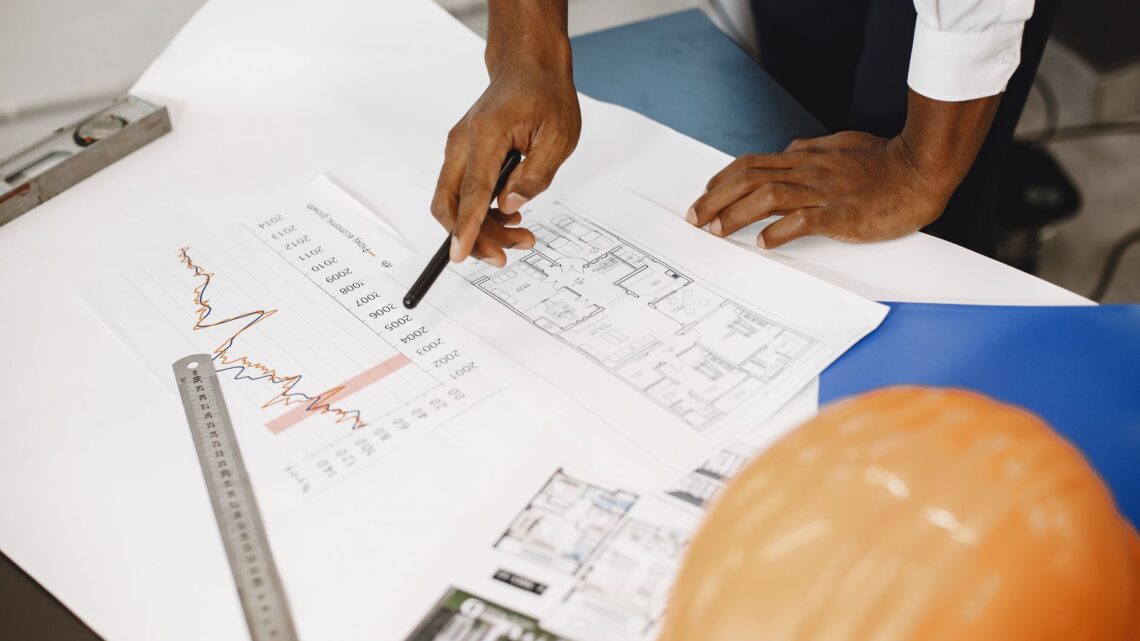 Hands of an architect analyzing blueprints and financial graphs at a desk.