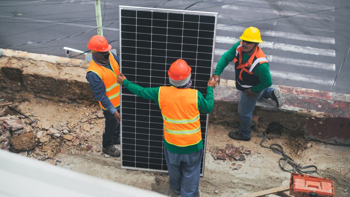 Workers wearing PPE install solar panels for sustainable energy at a construction site.
