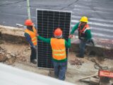Workers wearing PPE install solar panels for sustainable energy at a construction site.