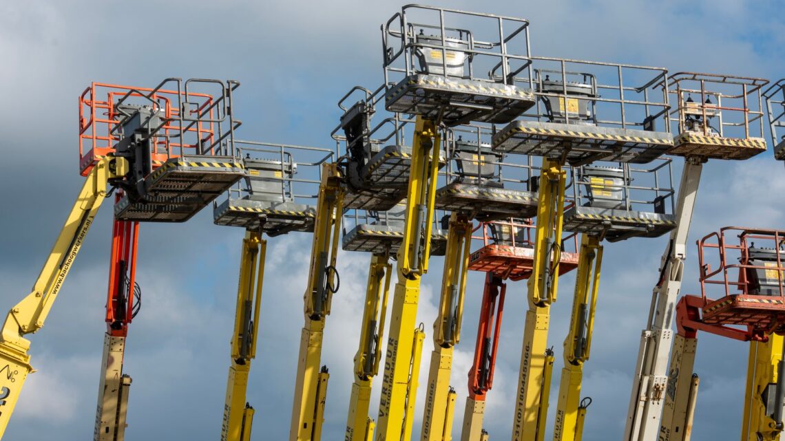 Multiple aerial work platforms with colorful booms reaching skyward against a cloud-dotted backdrop.
