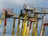 Multiple aerial work platforms with colorful booms reaching skyward against a cloud-dotted backdrop.