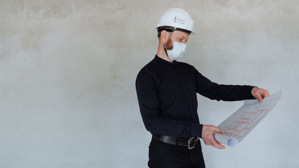 Architect in safety helmet and mask reviewing building plans indoors.