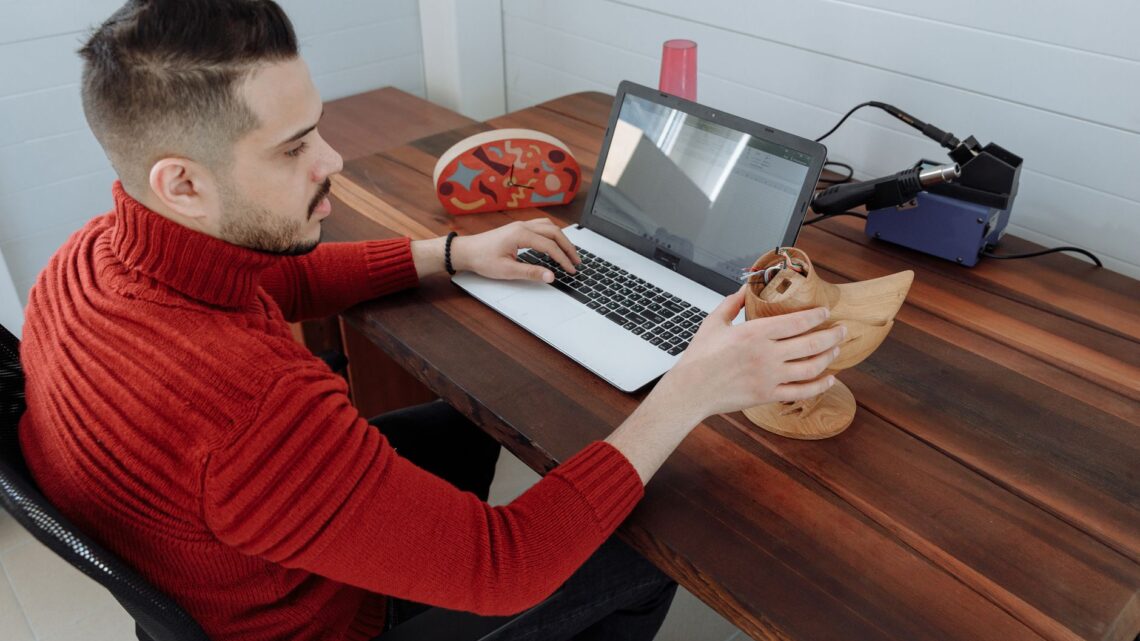 Engineer working on a laptop at a wooden desk, holding a 3D model in an office setting.