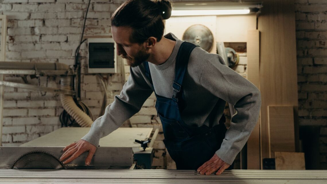 Man in denim overalls operating a circular saw in a woodwork shop.