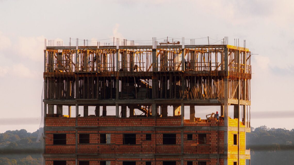Brick building under construction at sunset with workers visible on top floor.
