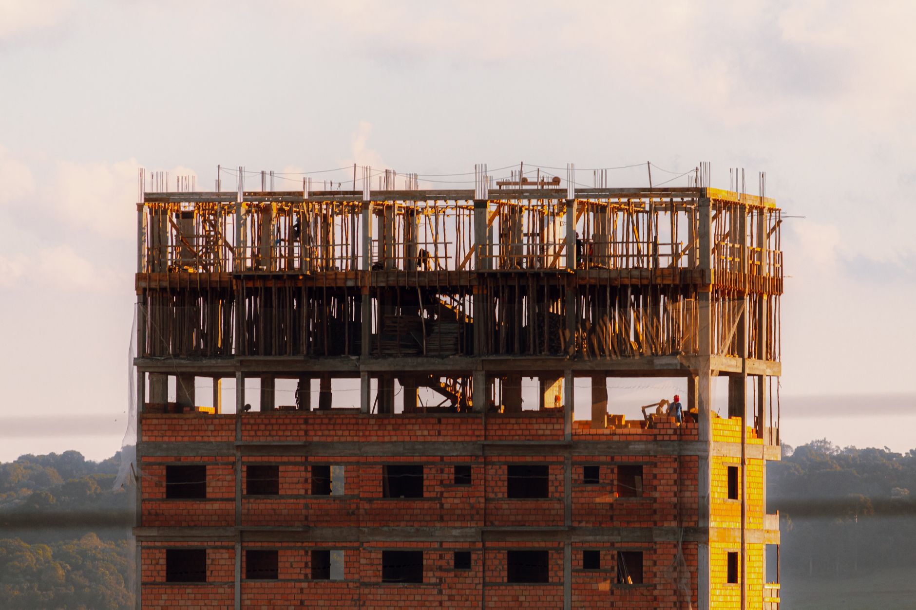 Brick building under construction at sunset with workers visible on top floor.